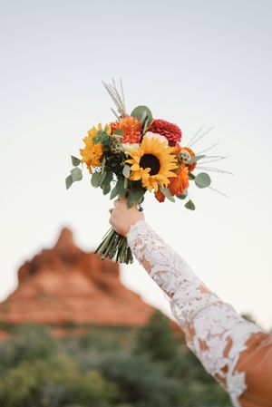 A lace-sleeved arm holds up a vibrant autumn bouquet with sunflowers against a desert mountain backdrop.