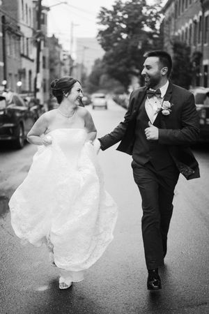 Black and white photo of a bride and groom joyfully running hand in hand down a city street on their wedding day.
