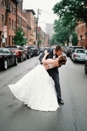Groom dips bride in a romantic kiss on a city street, her white lace wedding gown flowing, surrounded by parked cars.