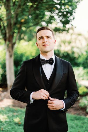 A series of wedding photos showing details of a black tuxedo and bow tie against a lush green outdoor background.