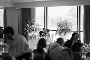Black and white photo of a wedding reception with floral arrangements by large windows and guests seated at tables.
