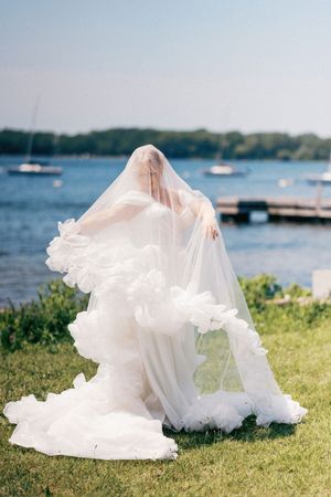 White wedding dress train flows elegantly across green grass with boats and water in the background at a lakeside venue.