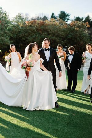 Wedding party in formal attire poses on green lawn with flowing white dresses creating elegant movement in sunlight.