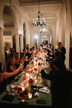 Elegant dinner party with long table set with candles and flowers under arched columns and chandeliers in dimly lit venue.