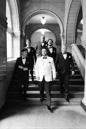 A black and white photo series showing groomsmen in tuxedos walking through an elegant arched hallway.
