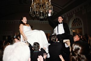 Wedding party members lift and carry a bride in her white gown during a celebratory reception moment.