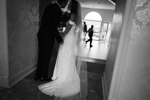 Black and white wedding photograph showing a couple embracing in a hallway while a silhouette appears in a distant doorway.