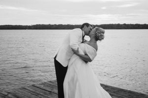 A romantic couple in white embraces on a wooden dock overlooking a serene lake at sunset.