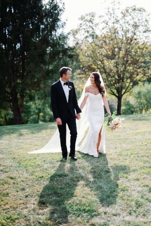 A couple in wedding attire walking together on a grassy hillside during golden hour.
