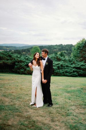 A couple in wedding attire shares romantic moments in a scenic meadow with rolling hills and evergreen trees in background.