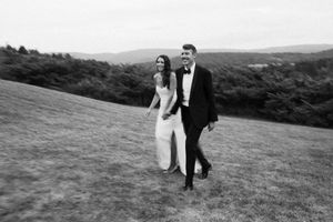 A couple in formal wedding attire poses on a scenic hillside overlooking a coastal landscape at sunset.