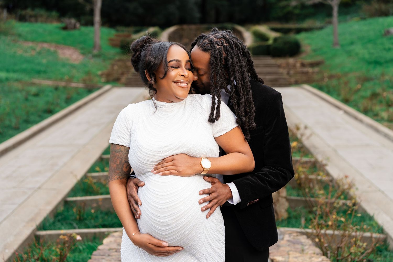 Sweet romantic moment captured outdoors as couple embraces on garden path with expectant mother wearing white dress.
