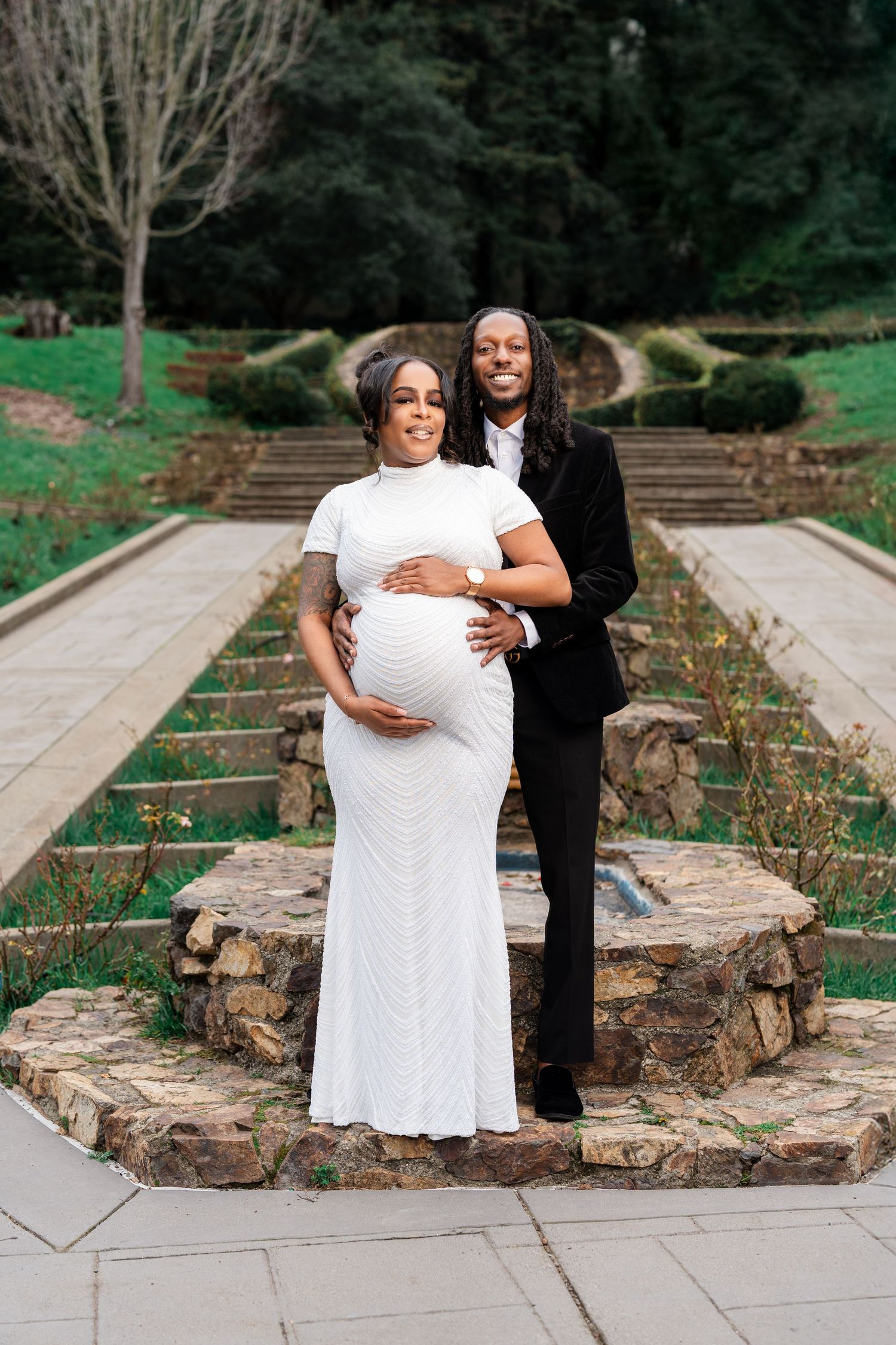 A couple poses on stone garden steps, with the expectant mother wearing a white gown and partner in black suit.