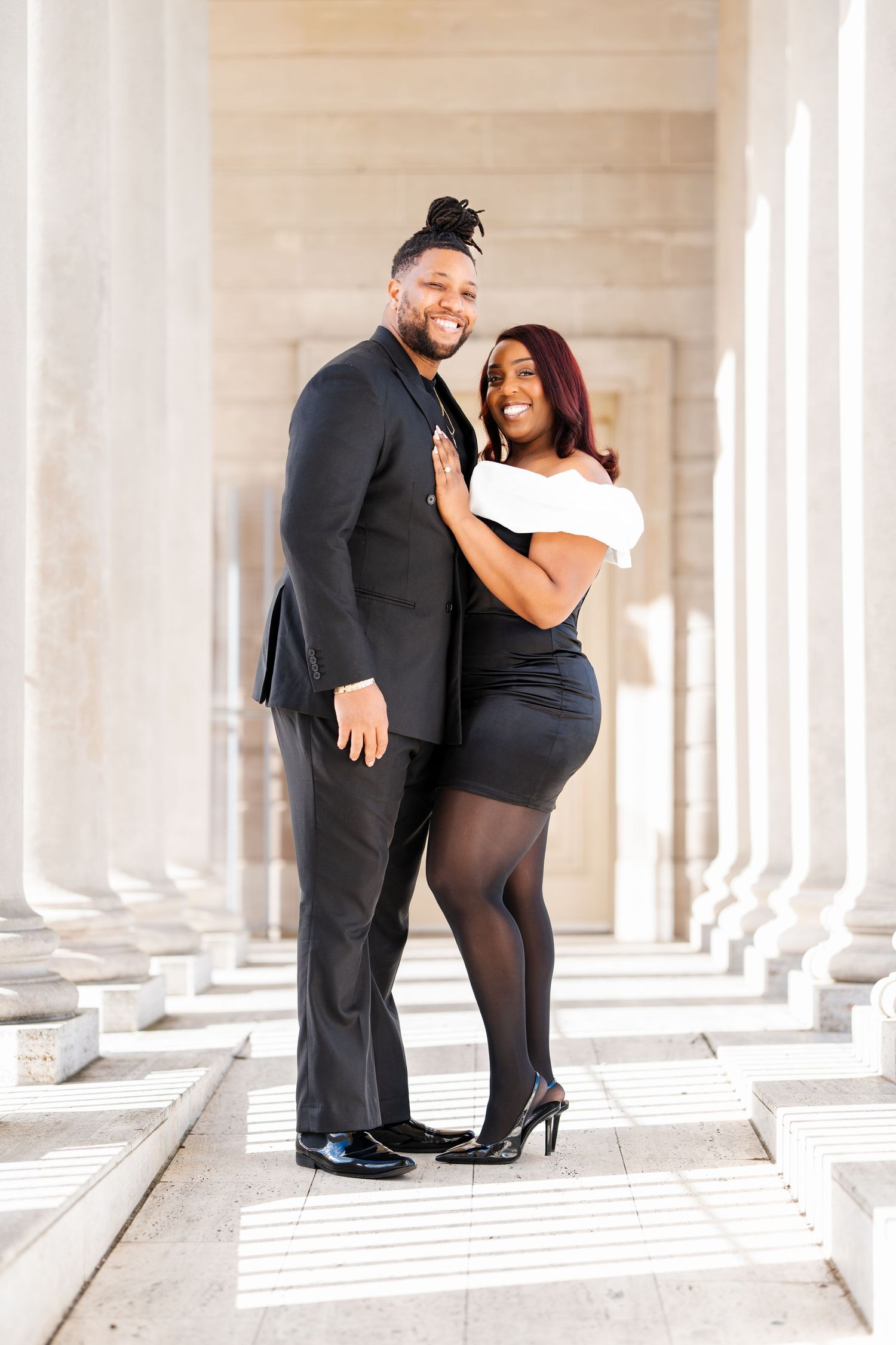 A couple poses together in formal attire between elegant white columns on marble steps.