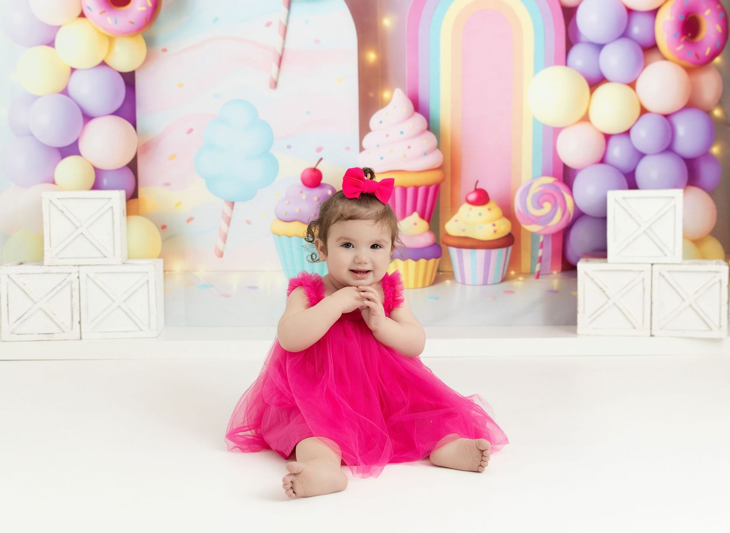 Toddler girl in pink tutu dress sits smiling before colorful balloon and cupcake birthday backdrop.