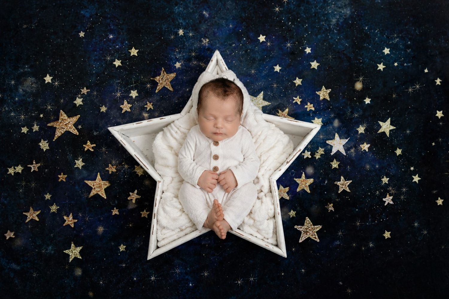 Newborn baby sleeping peacefully in a white star-shaped prop surrounded by gold stars on a dark blue background.