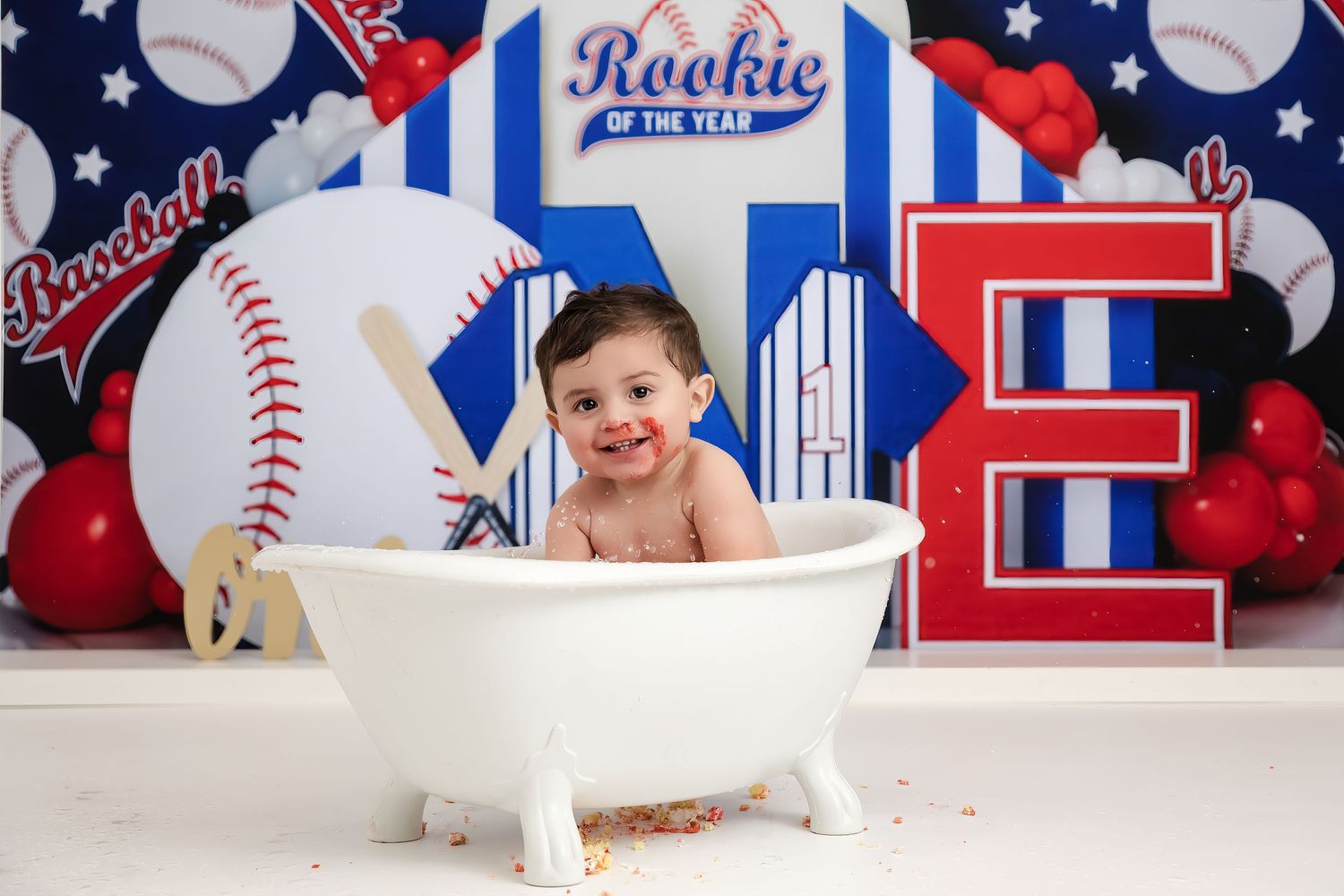 Baby boy sits in a white mini bathtub at a baseball-themed first birthday cake smash photo shoot.