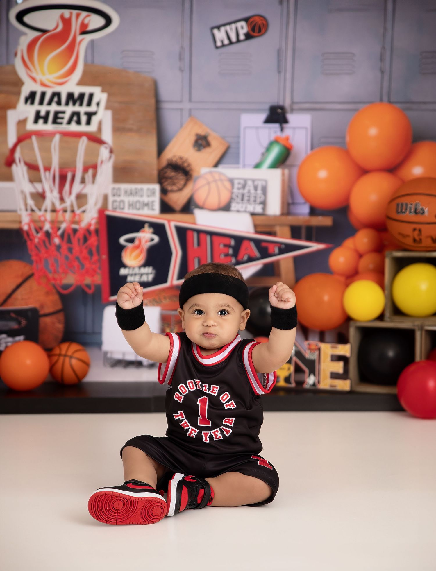 Baby in Miami Heat jersey sits with raised fists amid basketball-themed birthday party decorations.