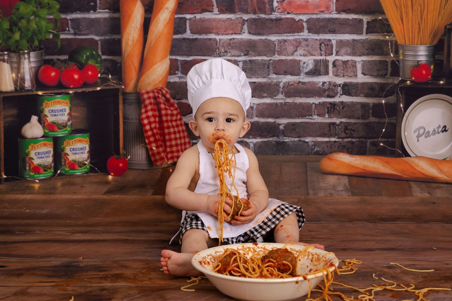 Cute baby in chef hat eating spaghetti from a large bowl in a kitchen-themed photo shoot.