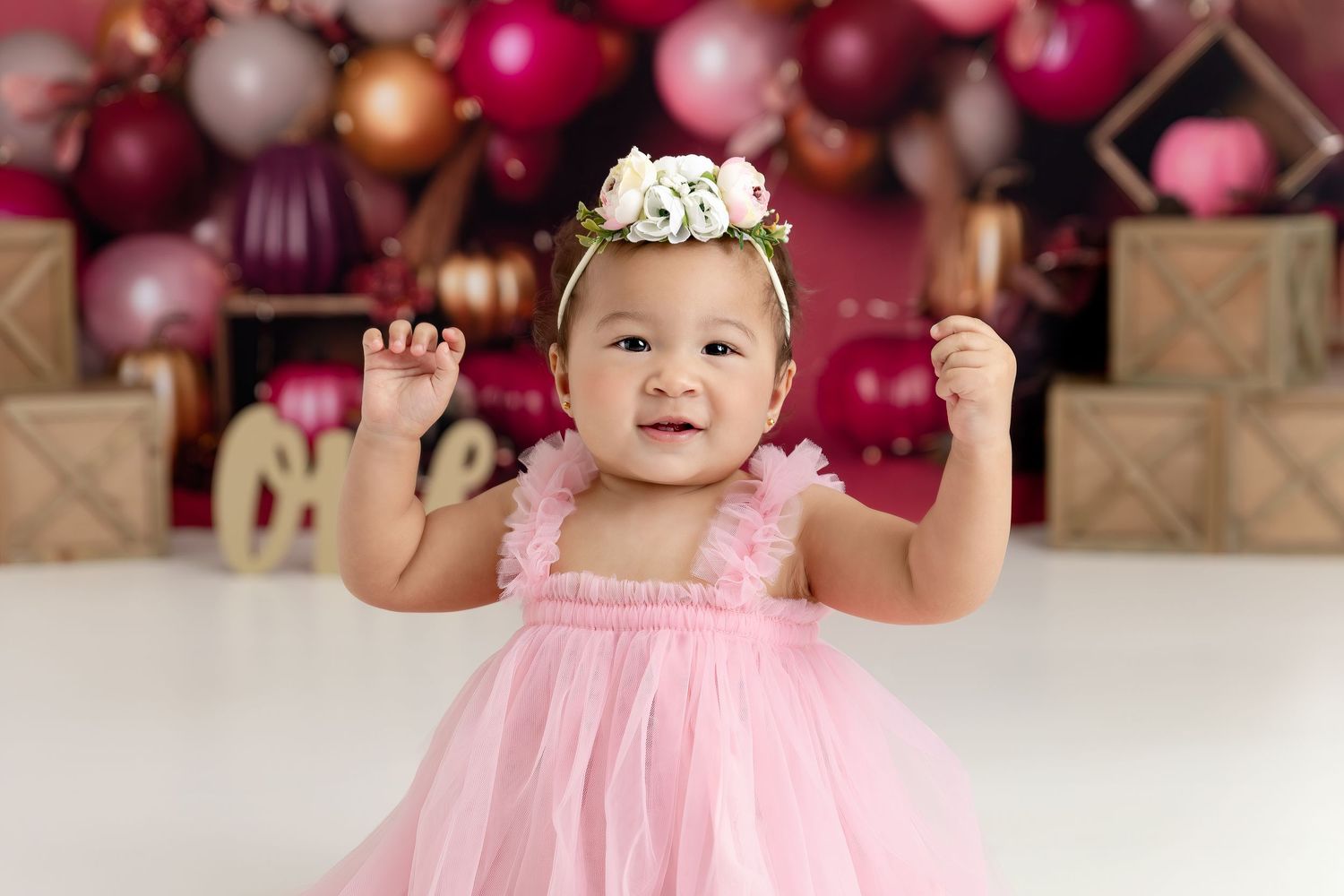 A smiling baby girl in a pink tulle dress and floral crown poses in front of colorful balloons and gift boxes.