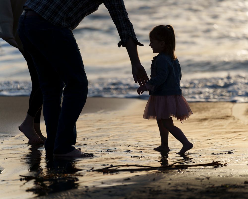 Silhouettes of people walking along a beach shoreline at sunset with waves and reflections in the wet sand.