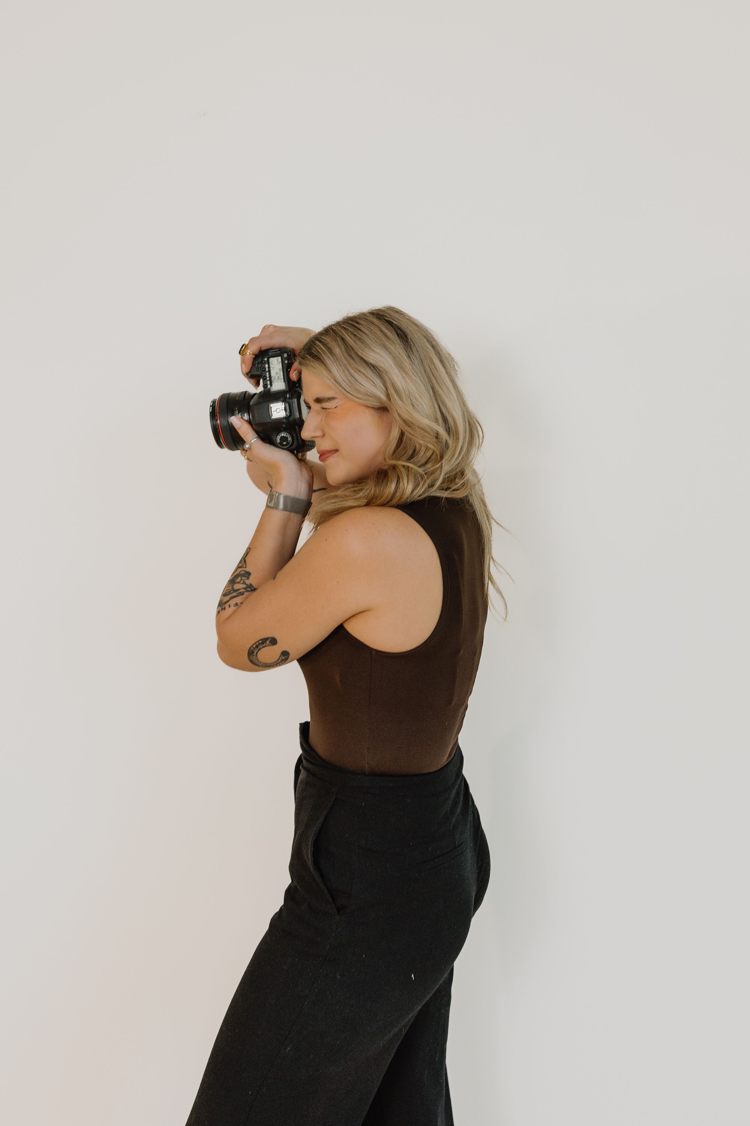 Blonde tattooed woman in brown top aiming film camera against white wall in side profile pose.