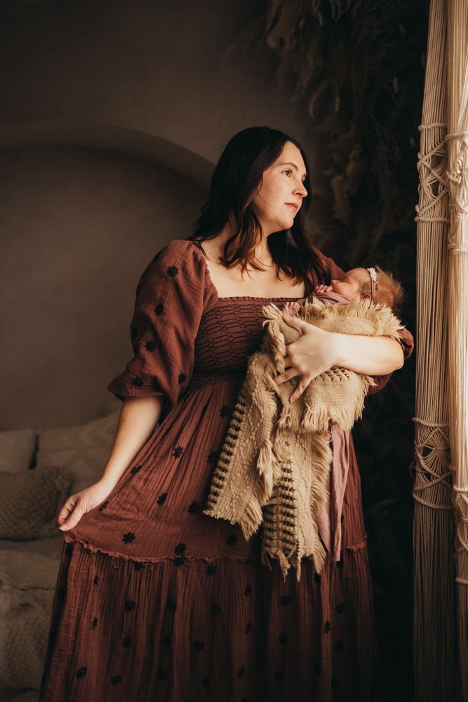 A mother in a burgundy dress tenderly holds her newborn baby near a window in a warmly decorated room.