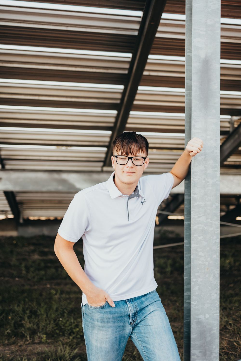 Young man in white polo shirt and jeans leaning against metal pole under corrugated roof structure.