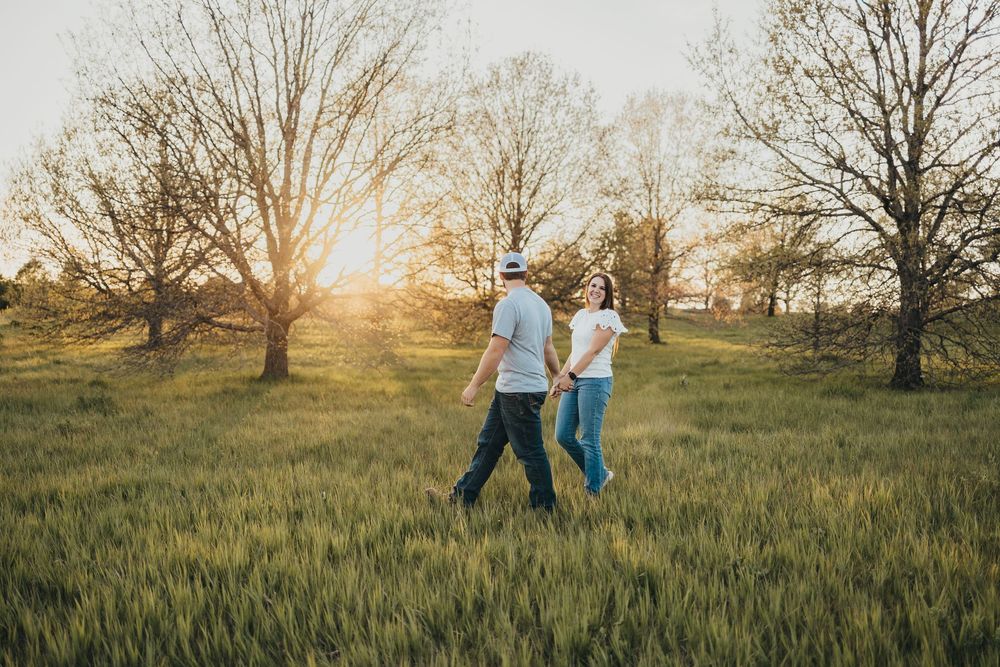 Couple walking hand in hand through a lush green field at golden sunset with bare trees in the background.