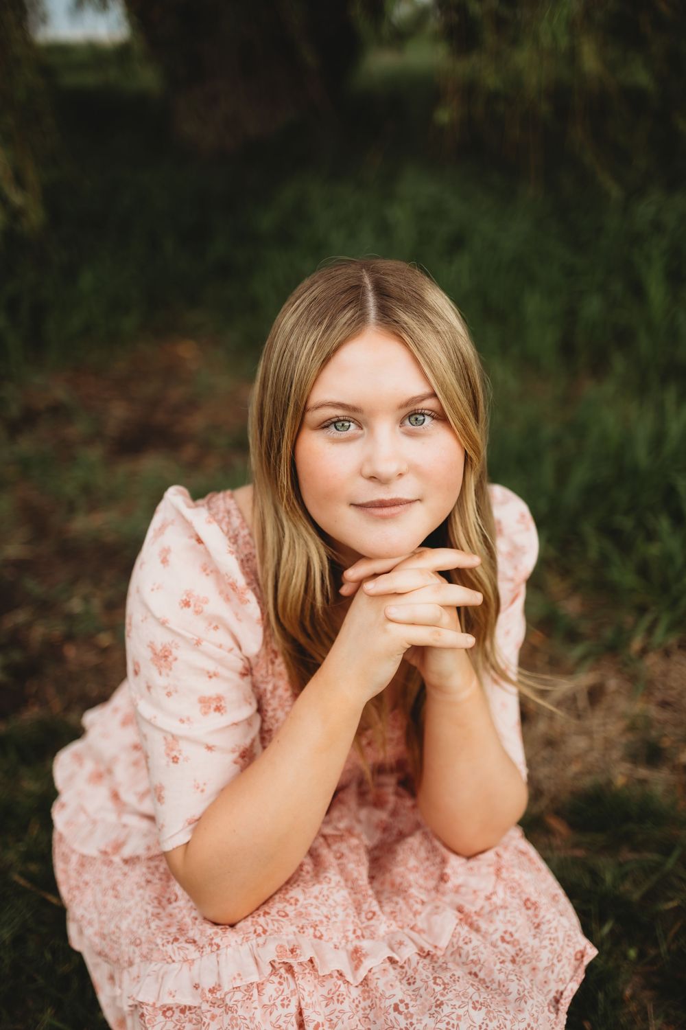 Young blonde woman in pink floral dress sitting outdoors, resting chin on hands, smiling softly at camera.