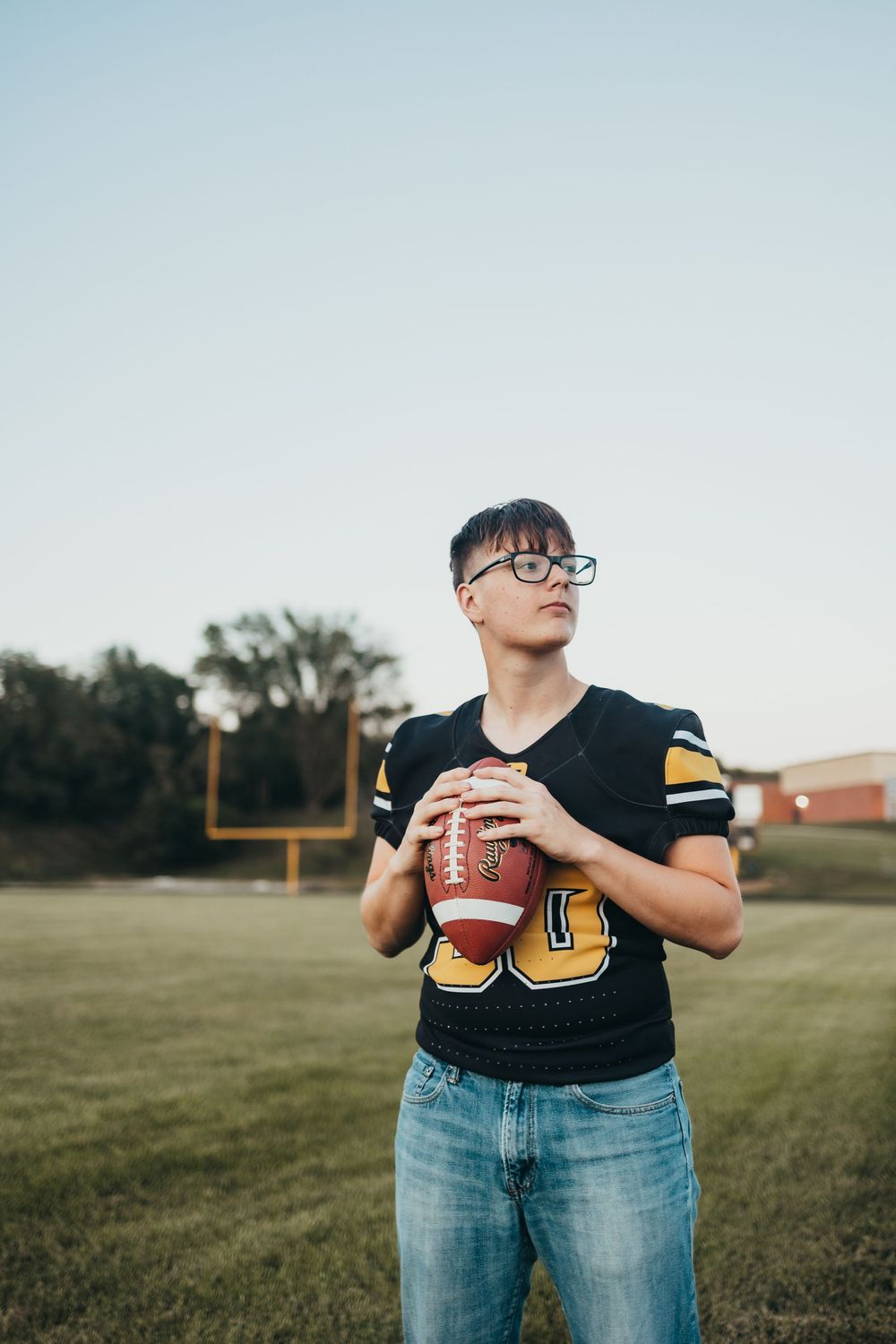 Young male football player in black jersey holding a football on a field with goalposts in the background.