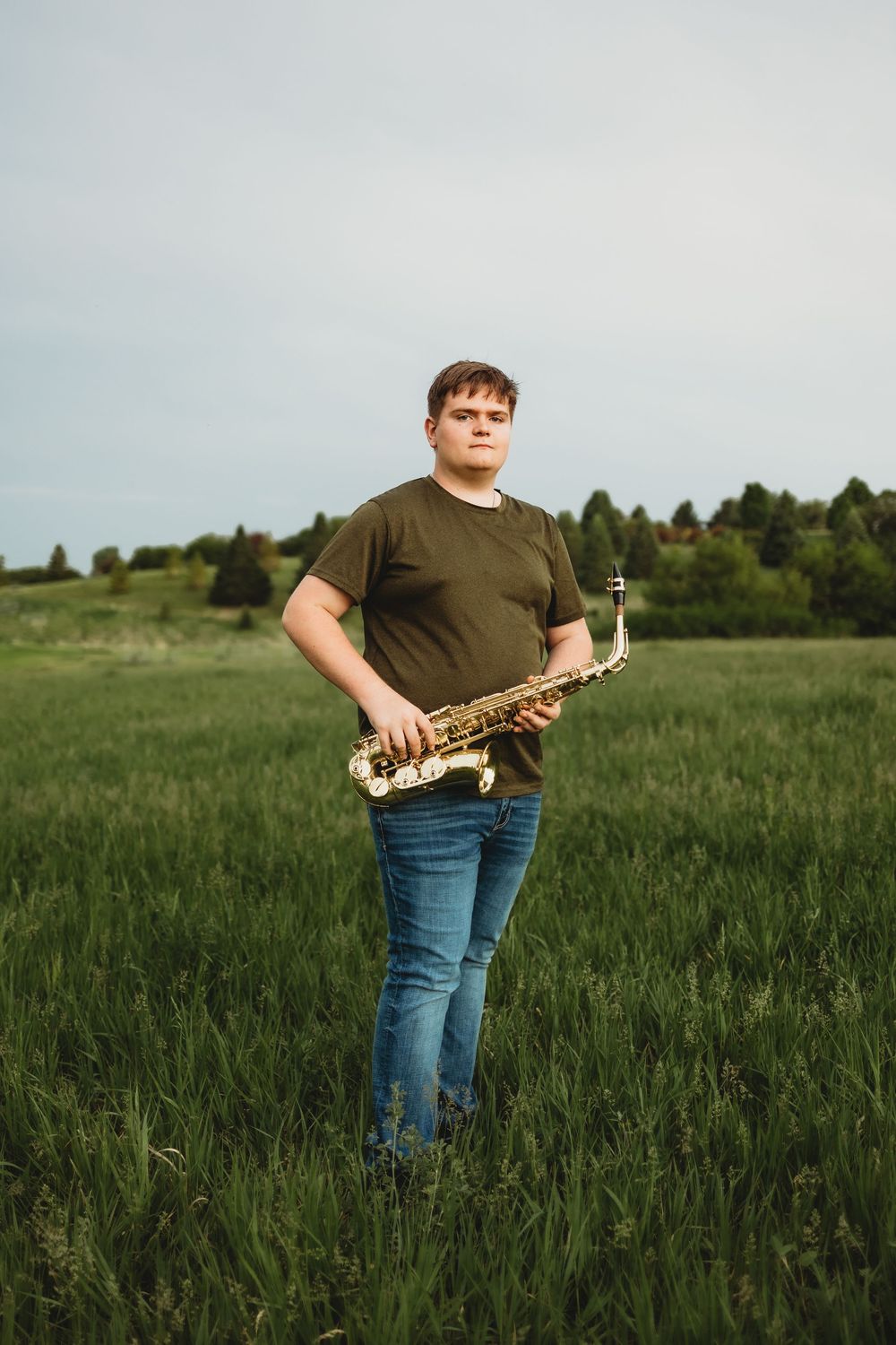Young man holding a saxophone while standing in a lush green field with trees in the background.