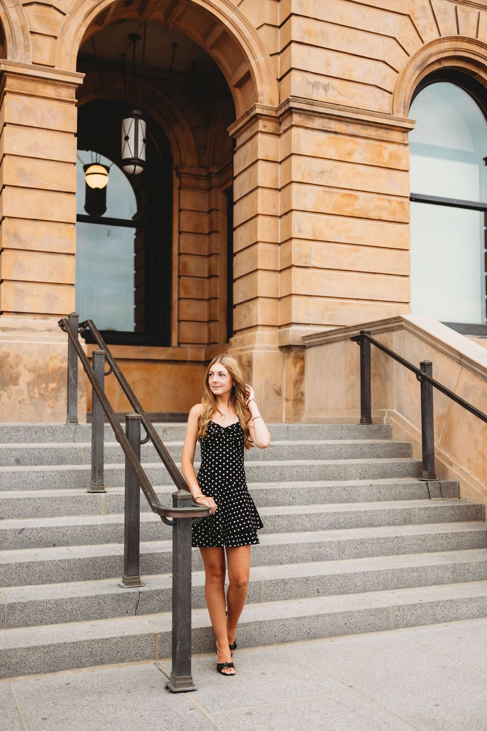 Young woman in black polka dot dress posing on stone steps of historic building with arched entrance.