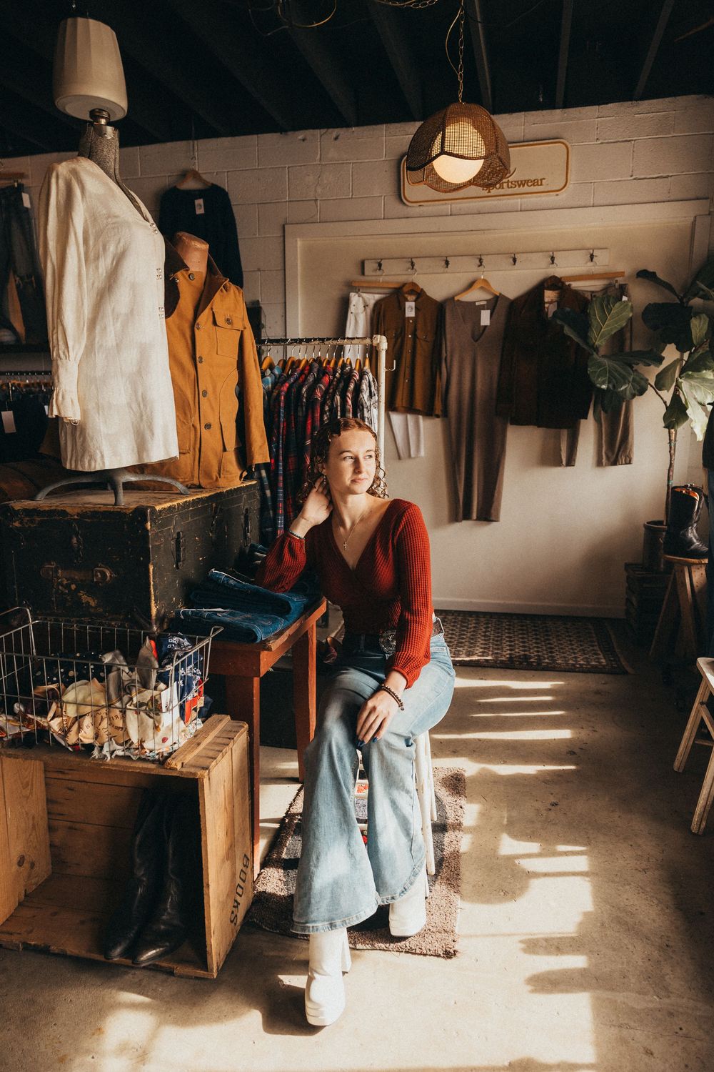 Woman in red sweater and jeans sits in sunlit boutique clothing store surrounded by hanging garments and rustic decor.