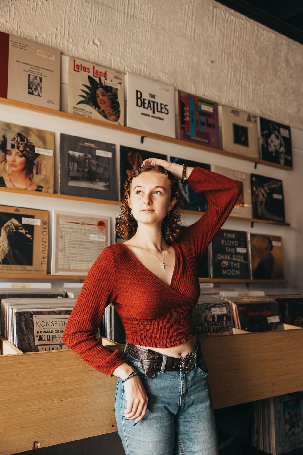 Young woman in red crop top and jeans poses in a vintage record store with Beatles and Pink Floyd albums on display.