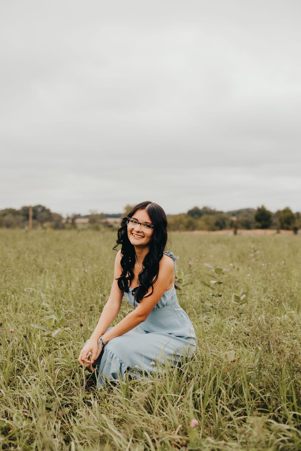 Young woman in a light blue dress smiling in a green field, wearing glasses with long dark hair.