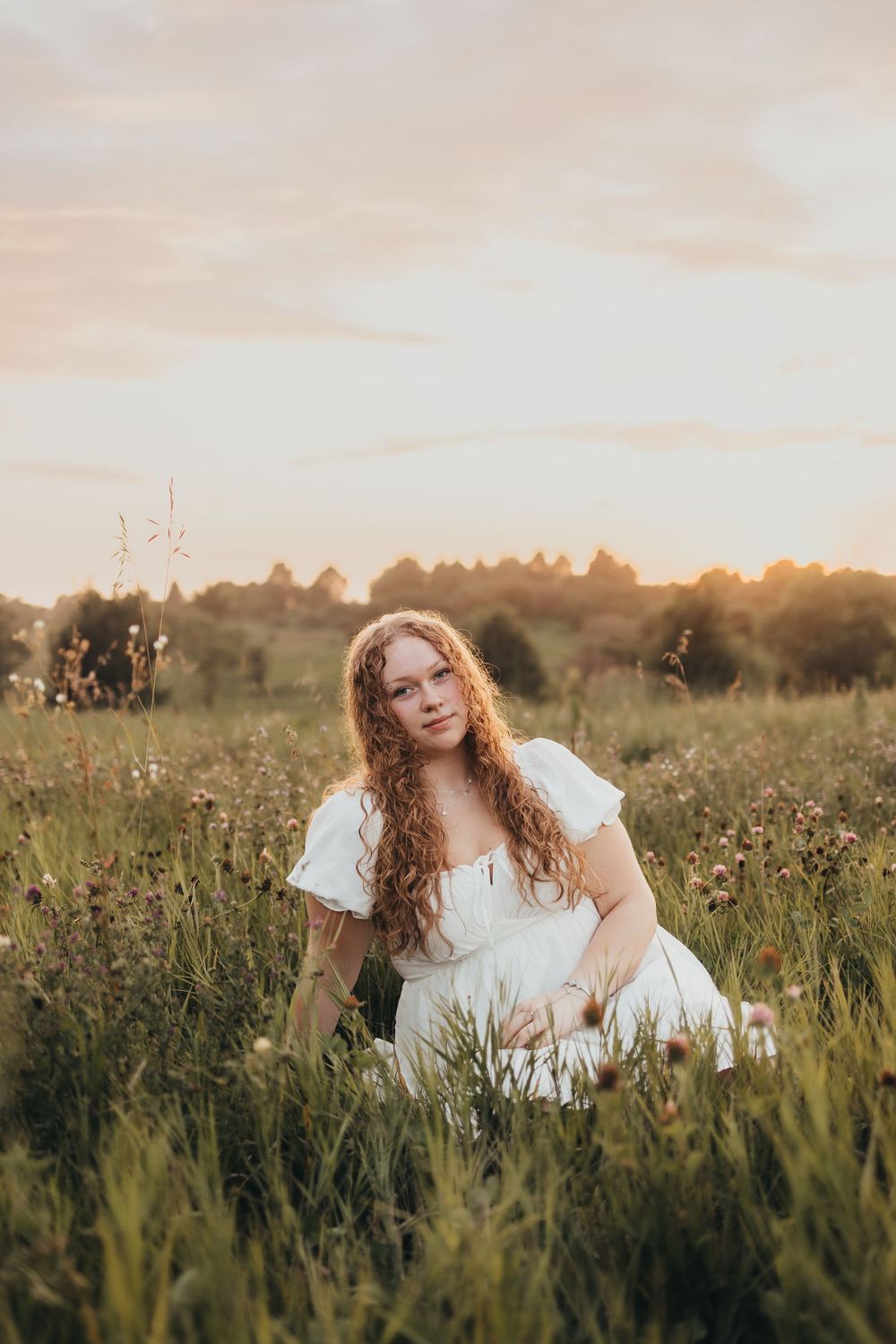Young woman with long curly red hair in white dress sitting in wildflower meadow at golden sunset.