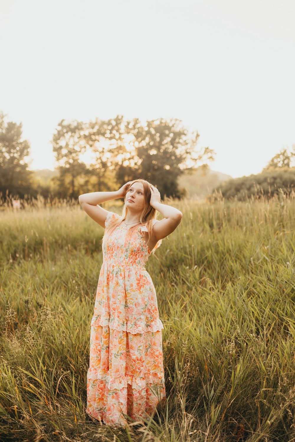 Woman in floral maxi dress standing in sunlit meadow with hands in hair, golden hour light filtering through trees.