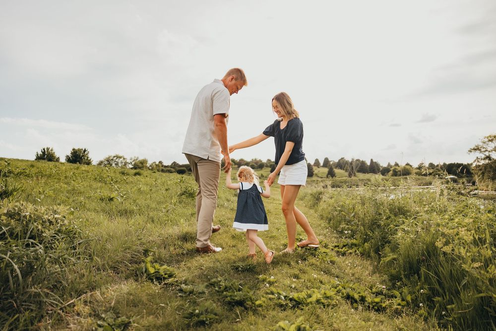 A young family holds hands with their toddler in a lush green field on a cloudy day.