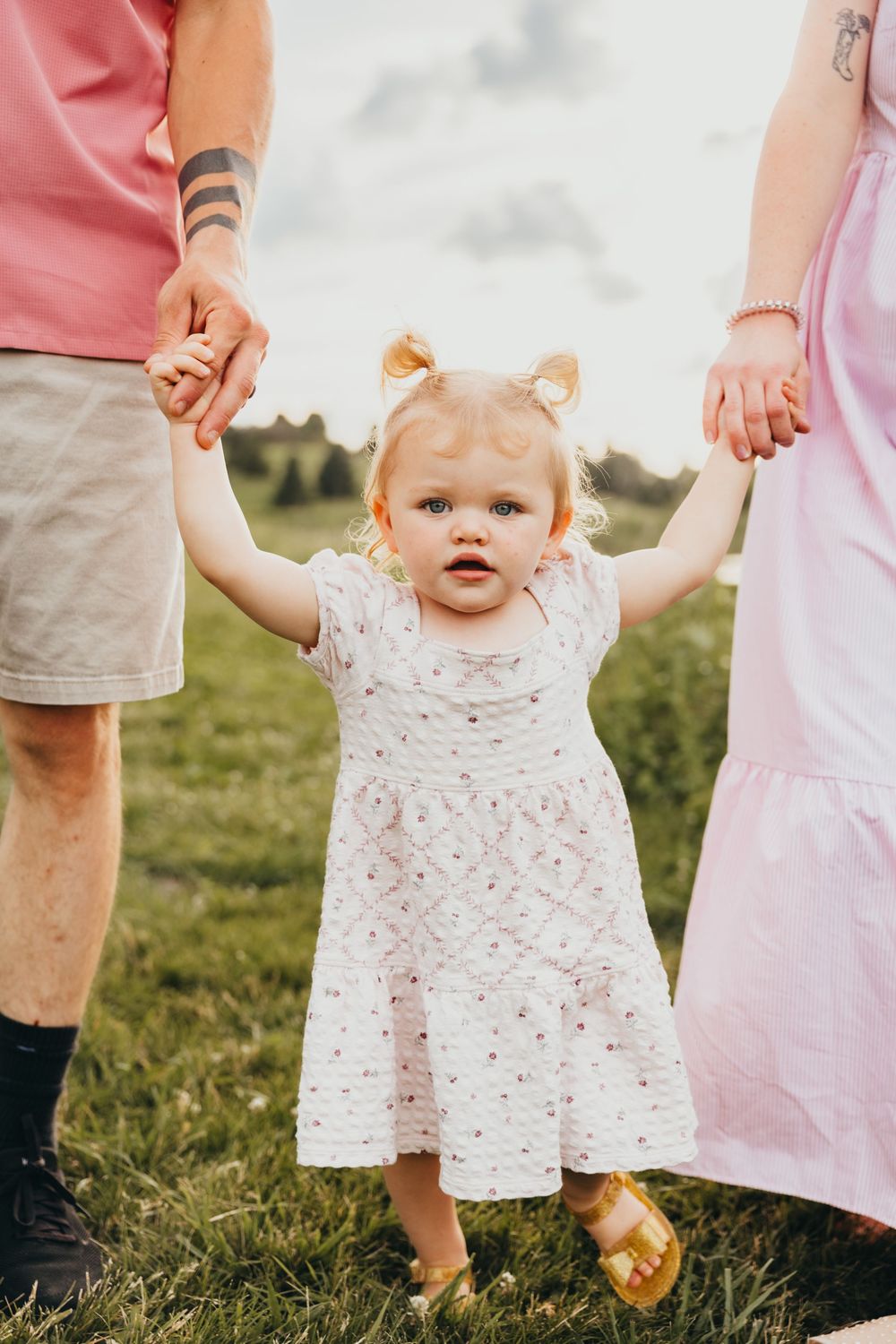 Toddler girl in white dress holds parents hands in green field.