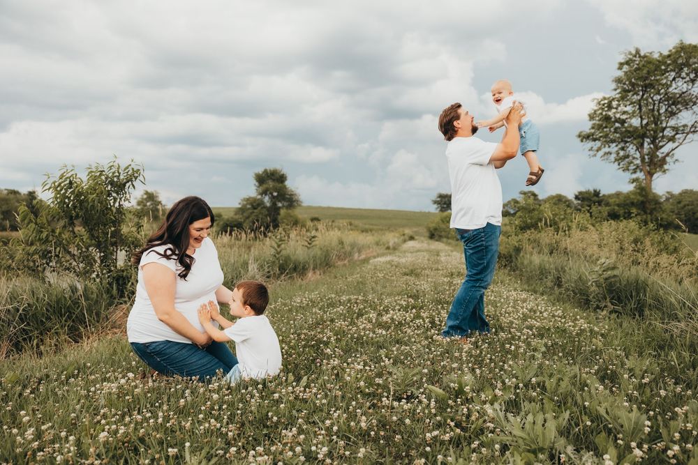 Family of four enjoying a sunny day in a wildflower field, with dad lifting baby while mom sits with toddler.