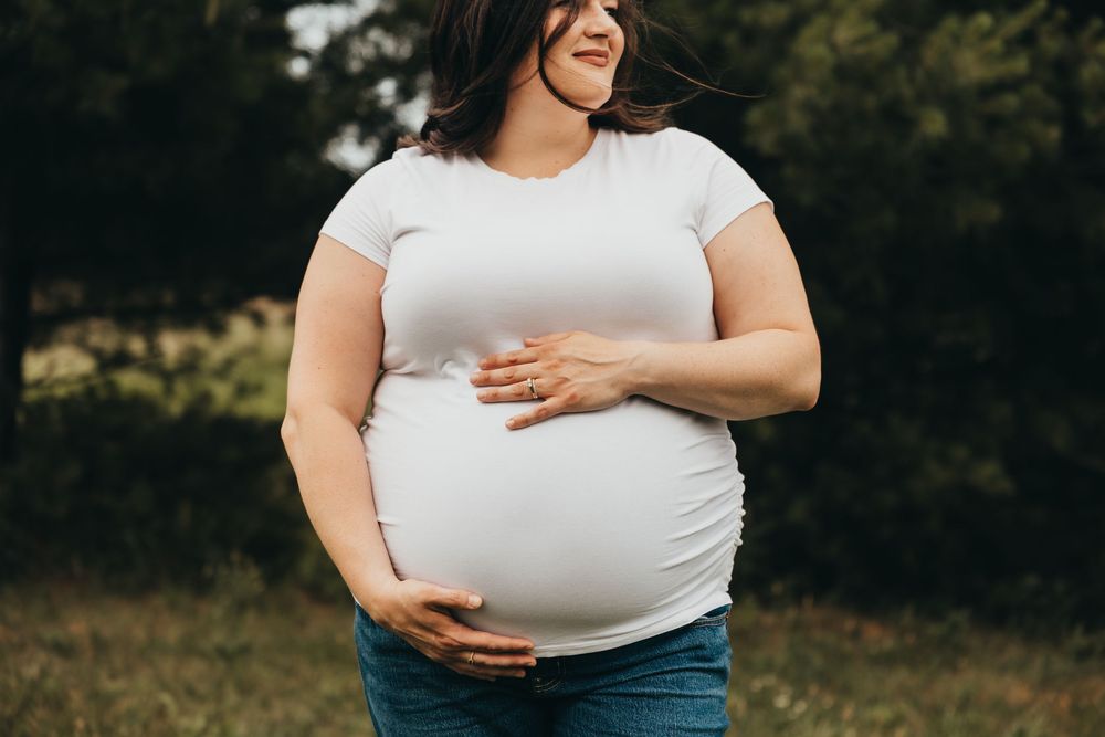 Pregnant woman in white t-shirt and jeans cradling her baby bump outdoors with green trees in background.