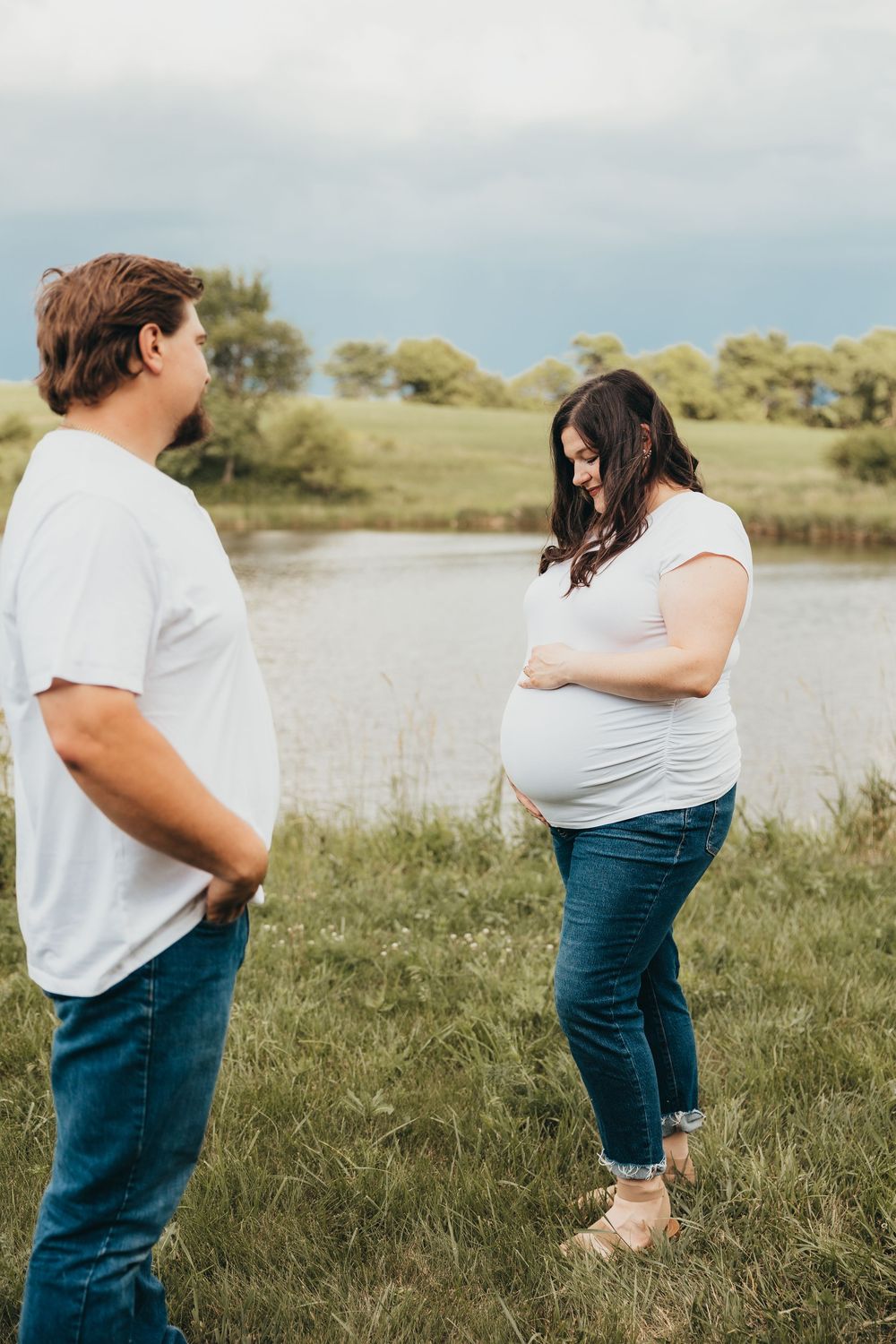 Pregnant woman in white tee and jeans holds her belly outdoors near a pond while her partner stands beside her.