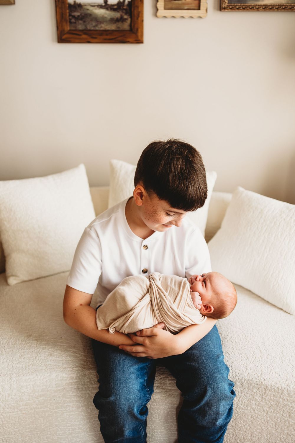 Young boy in white shirt and jeans gently holds swaddled newborn baby while sitting on a white bed.