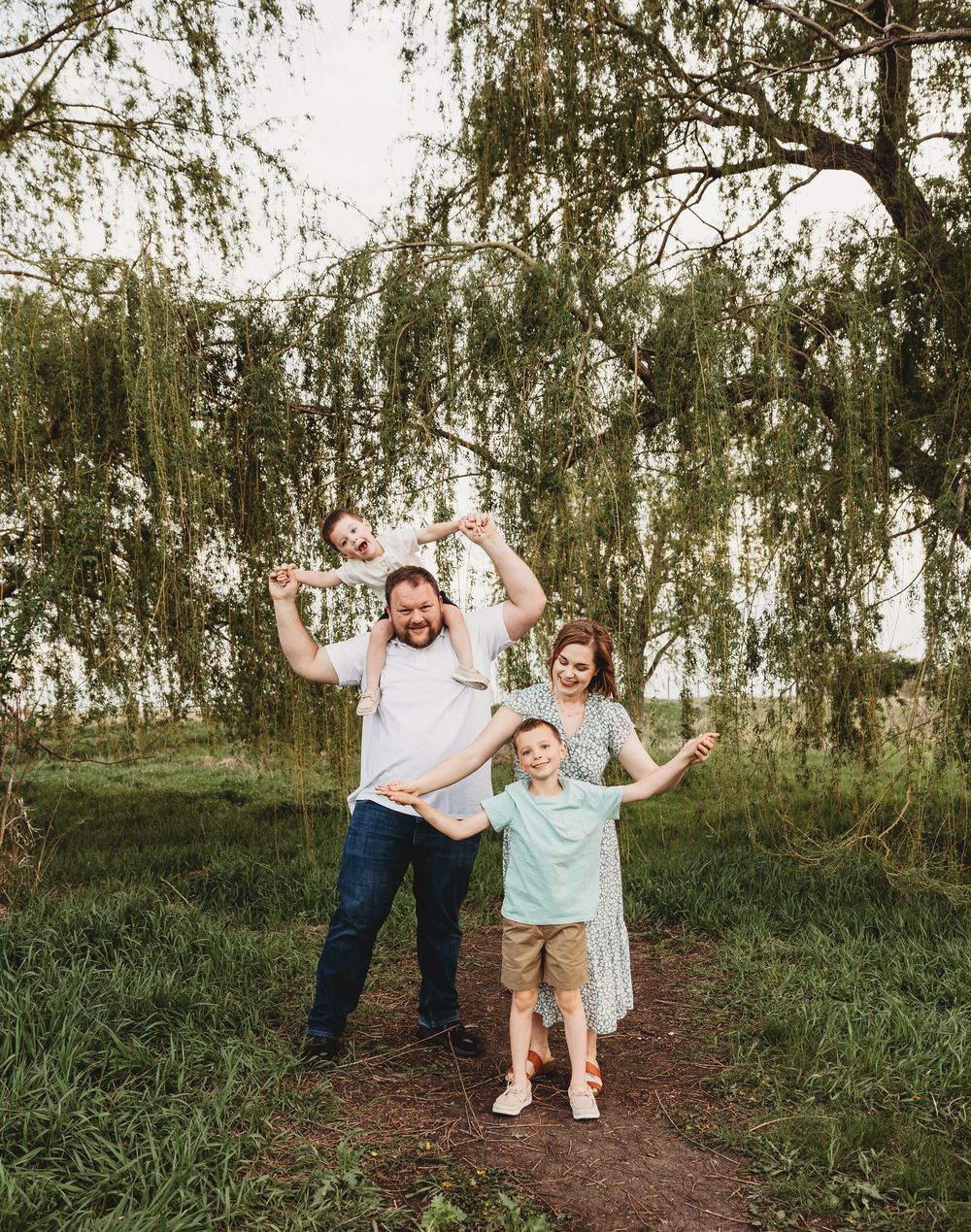 Happy family of four posing under a weeping willow tree, with child on dad's shoulders, outdoors in lush green setting.