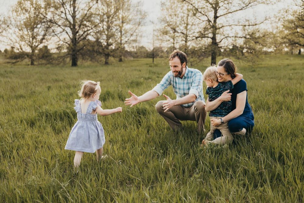 Family playing in green meadow, father reaching for toddler girl in blue dress while mother holds young boy at sunset.