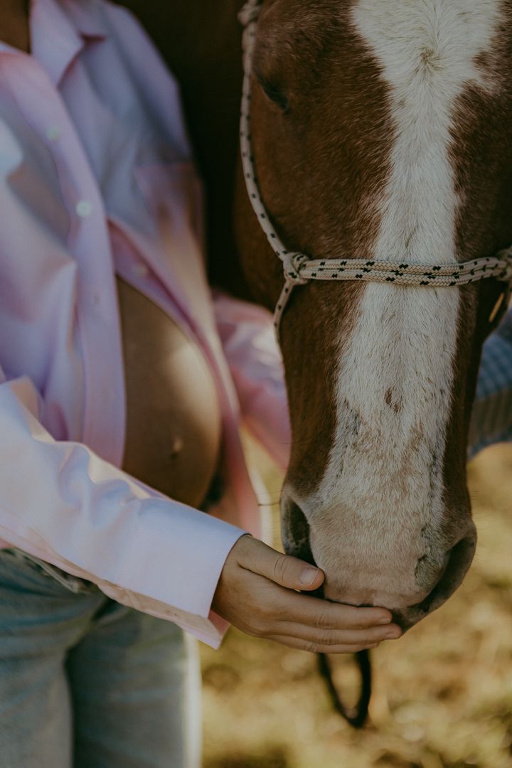 Pregnant woman in a pink shirt tenderly touching the nose of a horse in a field.