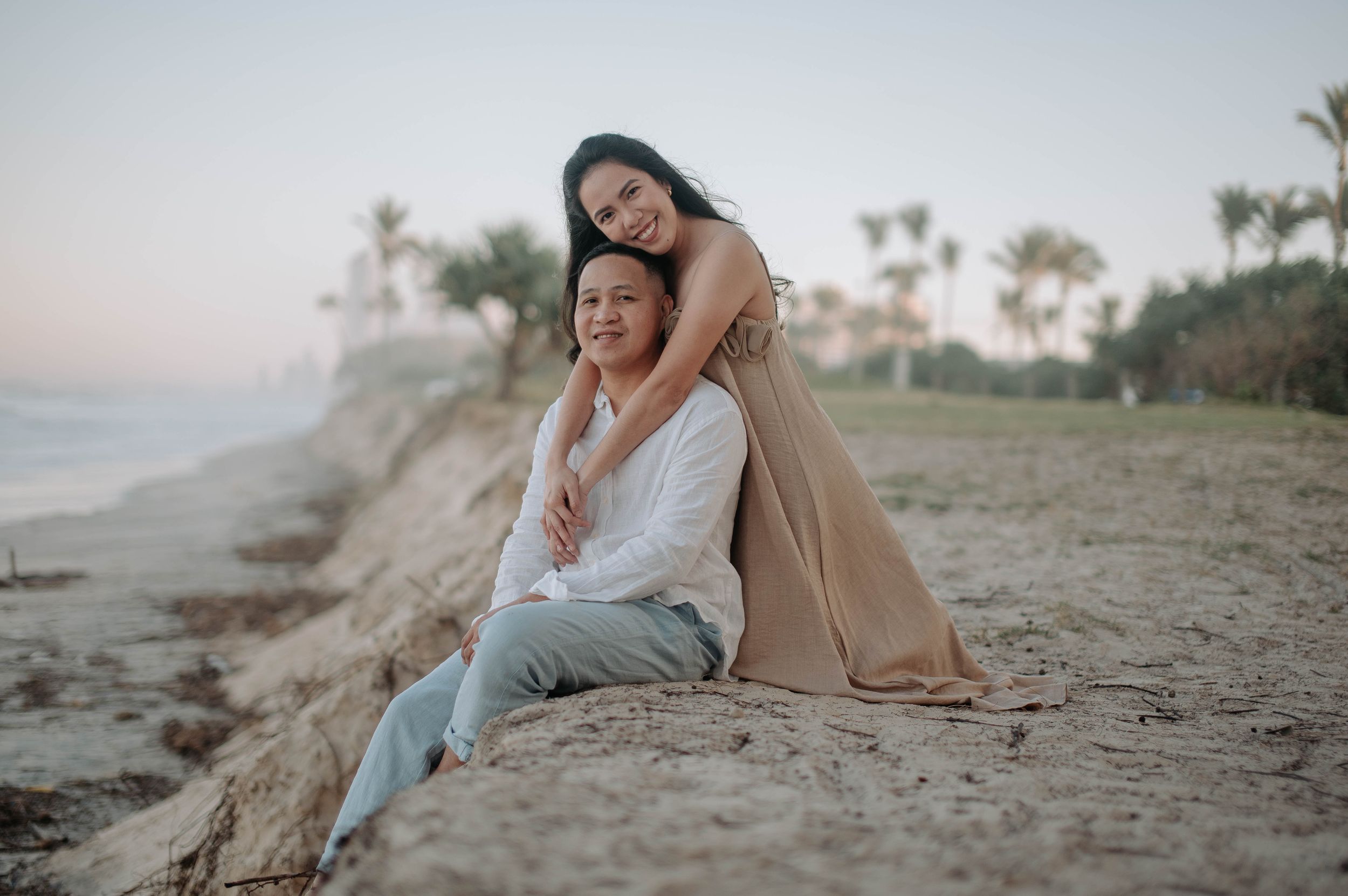 Couple posing on sandy beach at dusk, woman embracing man from behind, palm trees in soft background.