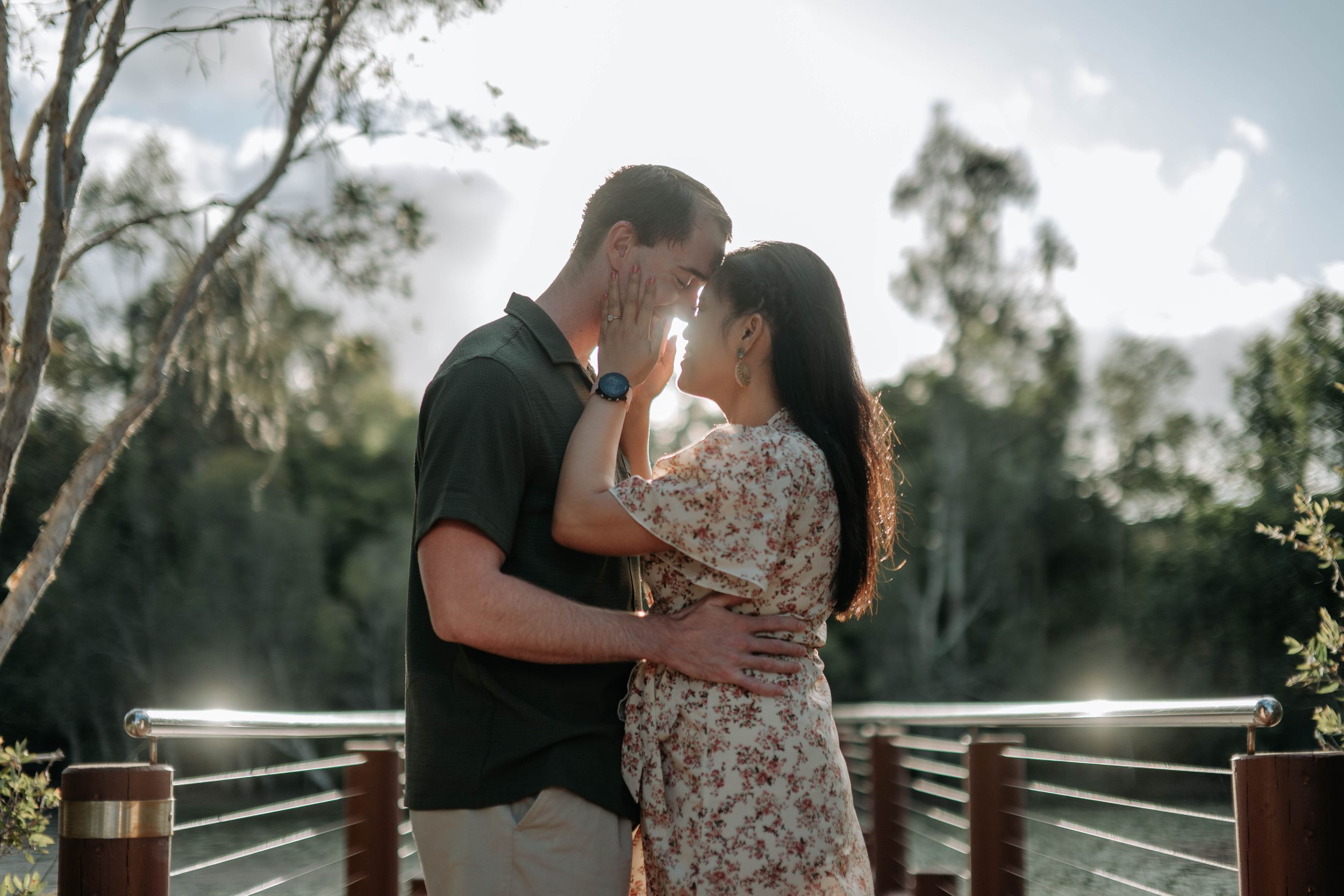 Couple sharing a romantic moment on a wooden bridge surrounded by lush green trees in soft natural sunlight.