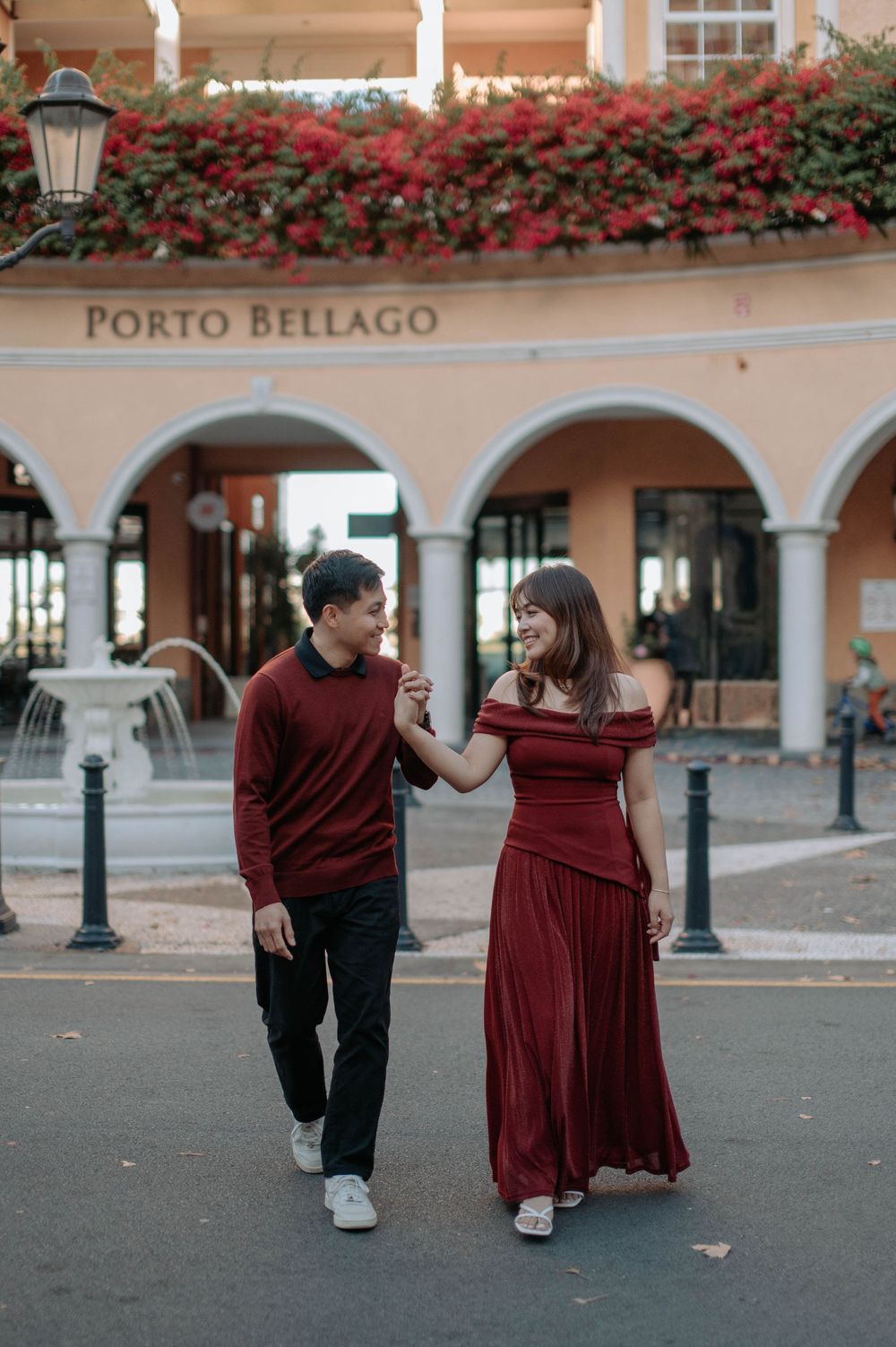 Couple in matching red outfits walking hand in hand in front of Porto Bellago building with arches and flowers.